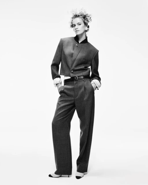 Blach-and-white full shot of a woman in a tailored suit with her hands in her pockets, striking a confident pose against a plain background.