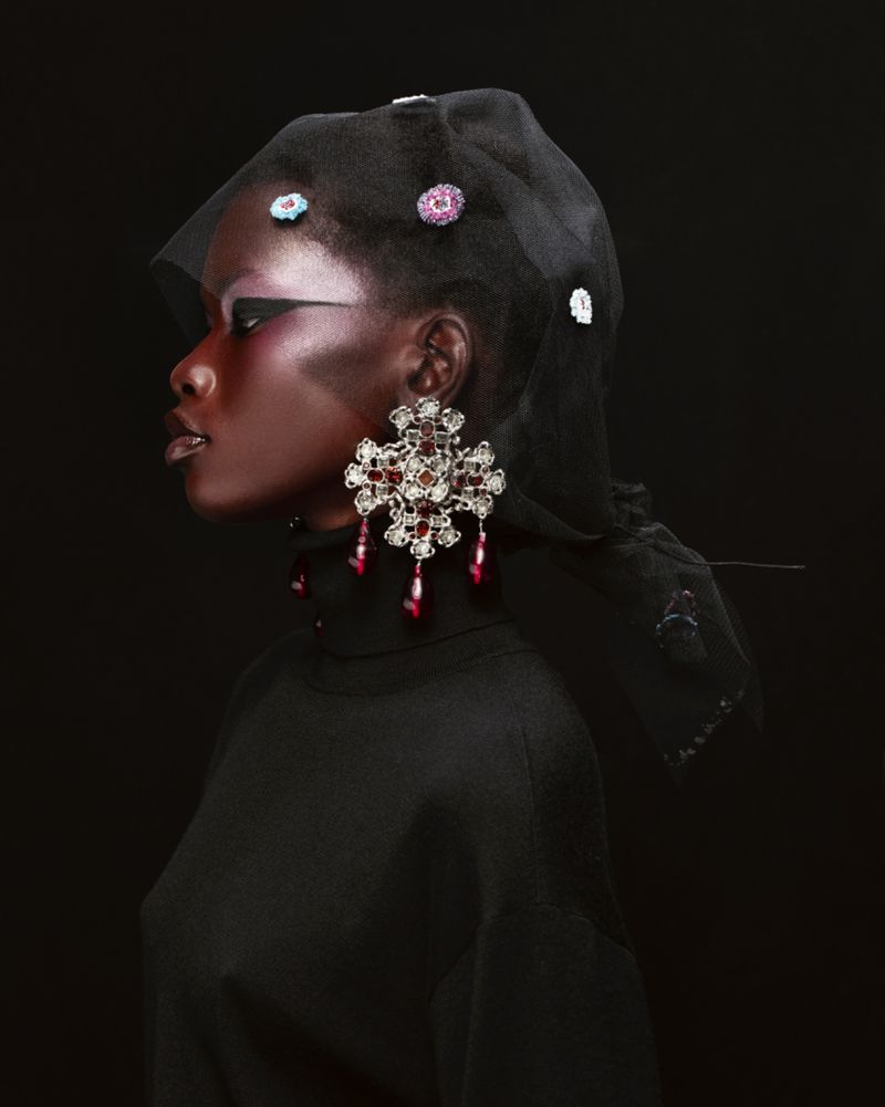 Profile shot of a dark-skinned model wearing a black turtleneck, a black sheer head covering with jewels, and large chandelier earrings.