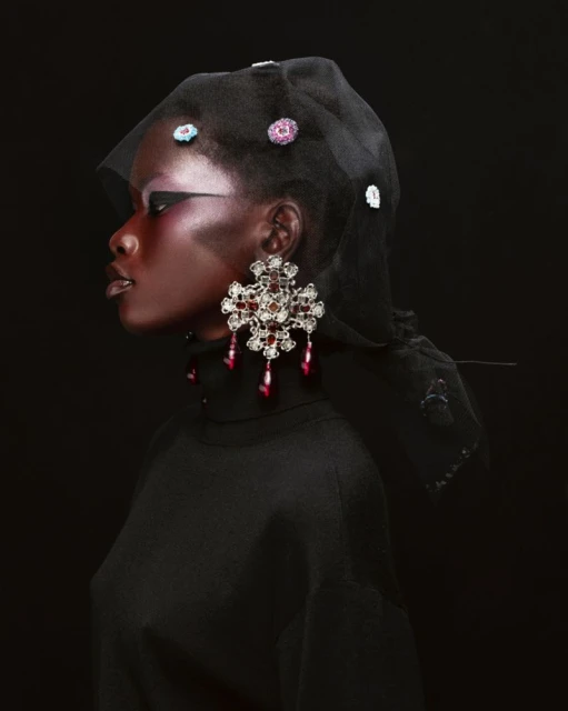 Profile shot of a dark-skinned model wearing a black turtleneck, a black sheer head covering with jewels, and large chandelier earrings.