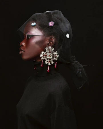 Profile shot of a dark-skinned model wearing a black turtleneck, a black sheer head covering with jewels, and large chandelier earrings.
