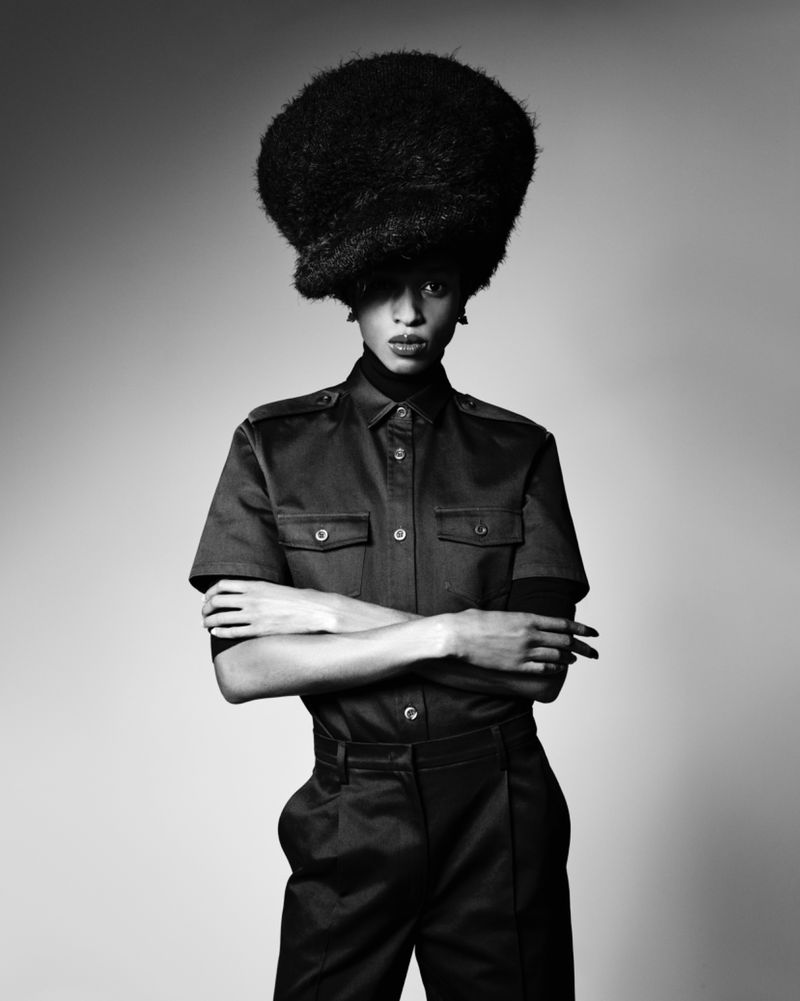 Model with a large voluminous textured hairstyle wearing a dark button-up shirt with crossed arms against a seamless grey backdrop.