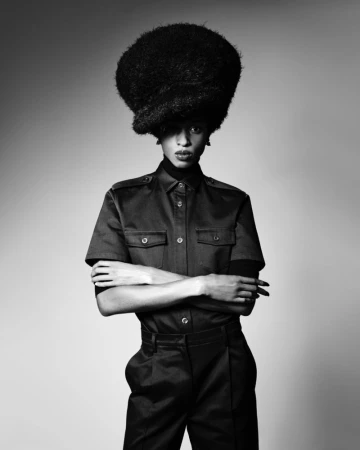 Model with a large voluminous textured hairstyle wearing a dark button-up shirt with crossed arms against a seamless grey backdrop.