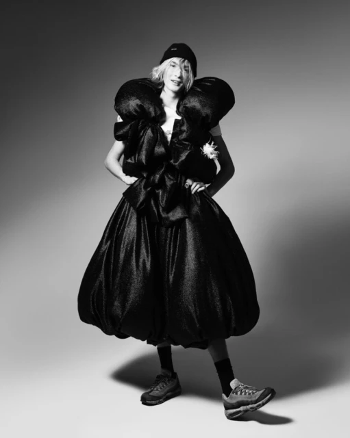 Adult wearing oversized avant-garde dress with black beanie and hiking shoes in a studio fashion shot with a neutral background.