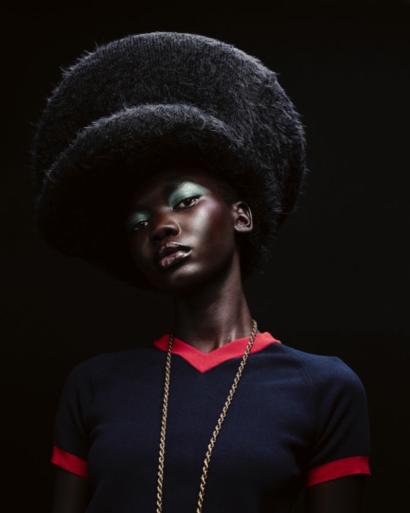 Striking portrait of a Black woman with green eyeshadow wearing a navy top, gold necklace, and oversized, fluffy black hat against black.