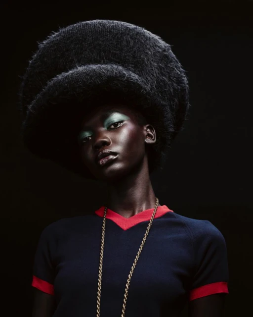 Striking portrait of a Black woman with green eyeshadow wearing a navy top, gold necklace, and oversized, fluffy black hat against black.