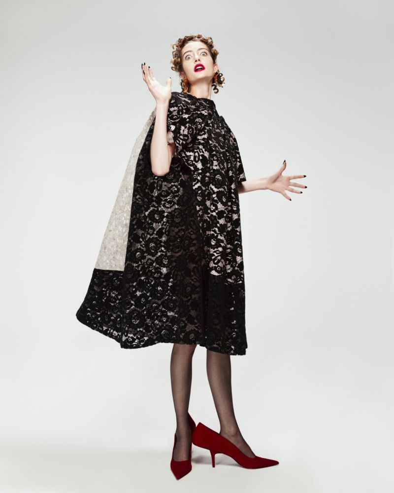 A woman in a black lace dress and red heels poses in a studio with arms raised, creating a fashion-forward and expressive visual.