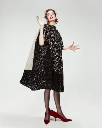 A woman in a black lace dress and red heels poses in a studio with arms raised, creating a fashion-forward and expressive visual.