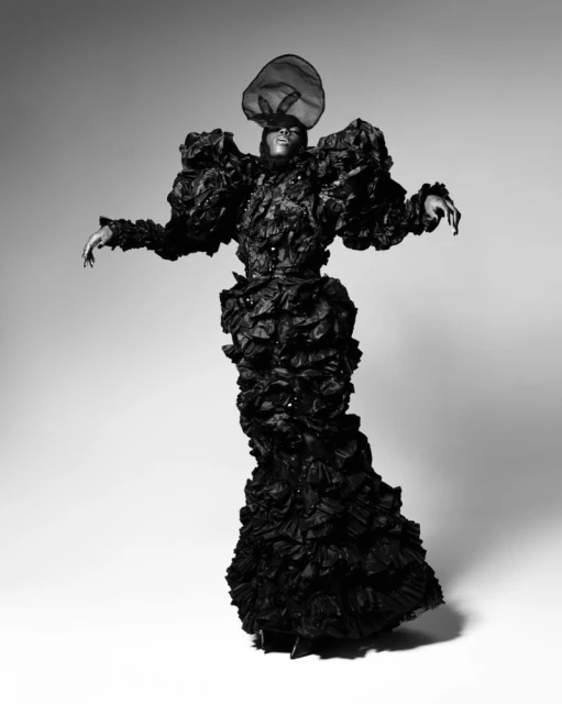 An African American model in a ruffled black haute couture gown and avant-garde headpiece stands in a graceful pose against a gray background.