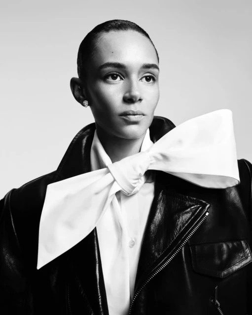 Elegant portrait of a woman with short hair, wearing a black leather jacket and large white bow tie against a neutral backdrop.