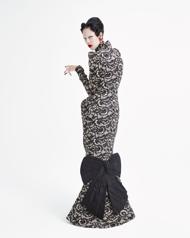 A woman in a lace gown stands in a white studio, looking over her shoulder with bright red lipstick and earrings. The dress has a large bow.