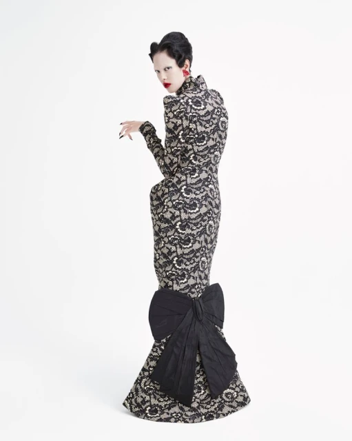 A woman in a lace gown stands in a white studio, looking over her shoulder with bright red lipstick and earrings. The dress has a large bow.