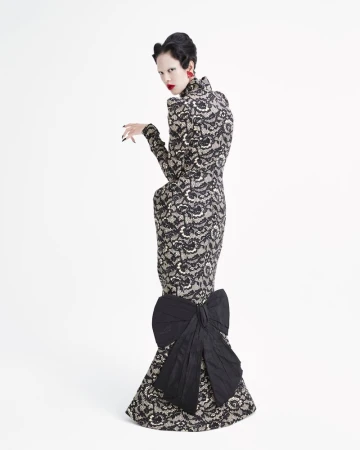 A woman in a lace gown stands in a white studio, looking over her shoulder with bright red lipstick and earrings. The dress has a large bow.