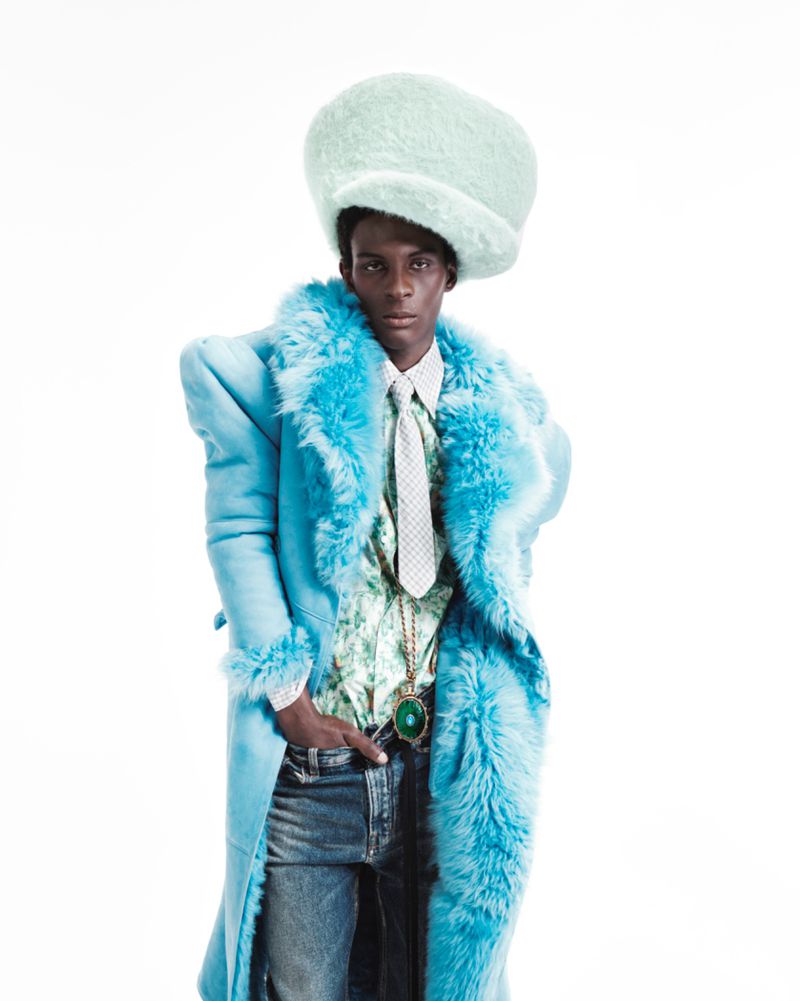 A person with dark skin poses in a blue fur-trimmed coat, jeans, and a mint green hat in a studio shot on a white background.