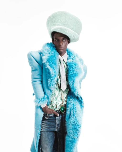 A person with dark skin poses in a blue fur-trimmed coat, jeans, and a mint green hat in a studio shot on a white background.