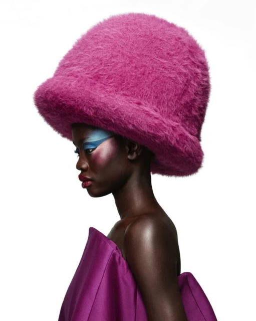 Profile portrait of a Black woman wearing a large magenta fuzzy hat, draped magenta fabric, and blue makeup against a white background.