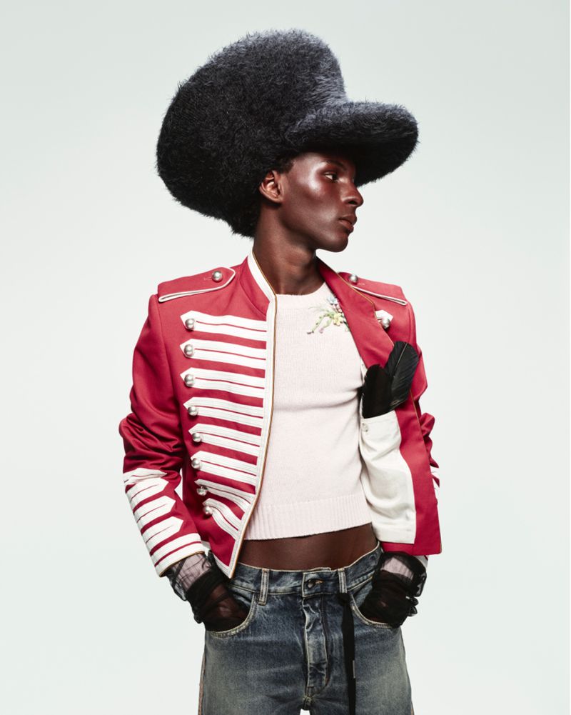 Fashionable dark-skinned man in a red military-style jacket and black hat posing in a studio. He also wears jeans and gloves.