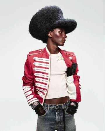 Fashionable dark-skinned man in a red military-style jacket and black hat posing in a studio. He also wears jeans and gloves.