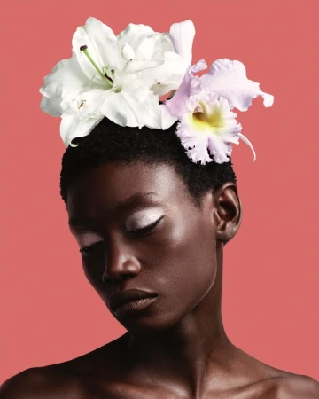 Portrait of a short-haired African-descent adult woman with closed eyes. She is adorned with white and light-purple flowers.