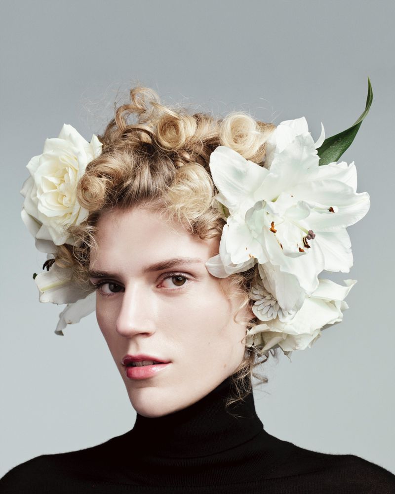 Close-up portrait of adult female with curly blonde updo adorned with large white flowers, wearing a black turtleneck against a light gray background.