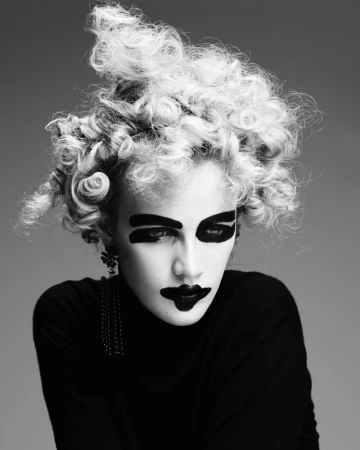 A blonde model with voluminous, tight curly hair and bold black face paint around eyes and lips, wearing a simple black turtleneck.