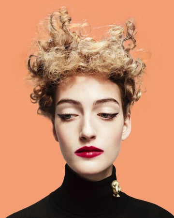 Close-up portrait of a woman with sculpted blonde curly hair, dark winged eyeliner, red lipstick, and a black turtleneck against a solid peach background.