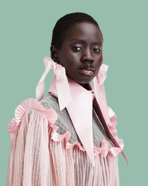 Close-up studio portrait of an African American woman wearing a pink ruffled top and light pink ribbon collar on a light green backdrop.