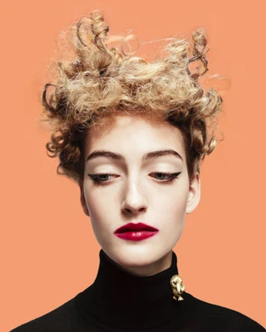 Close-up portrait of a woman with sculpted blonde curly hair, dark winged eyeliner, red lipstick, and a black turtleneck against a solid peach background.
