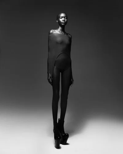 Model standing full-length in a dark fitted bodysuit against a neutral studio backdrop captured by Willy Vanderperre for Elle Belgium.