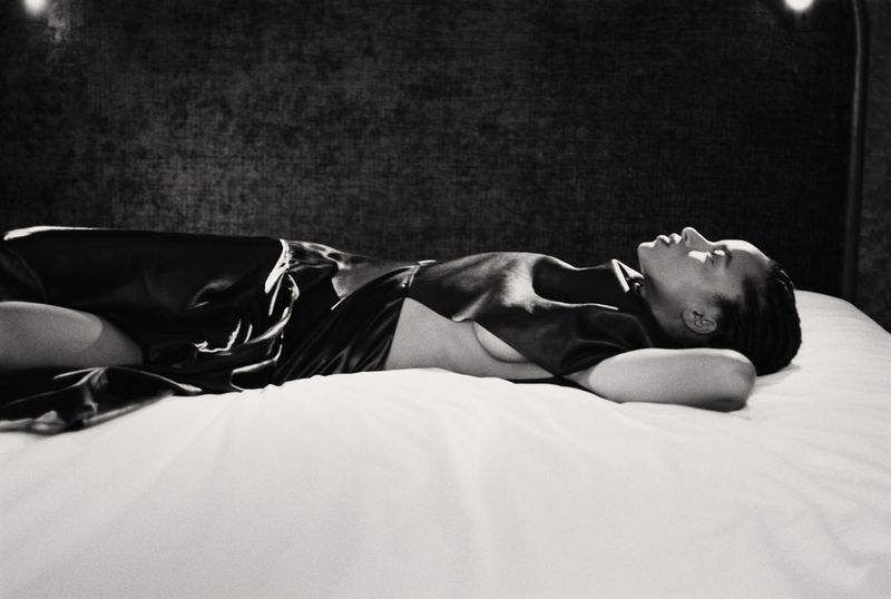 Monochrome studio portrait of a woman lying on a white bed in a dark dress, looking up toward a light, creating a tranquil scene.