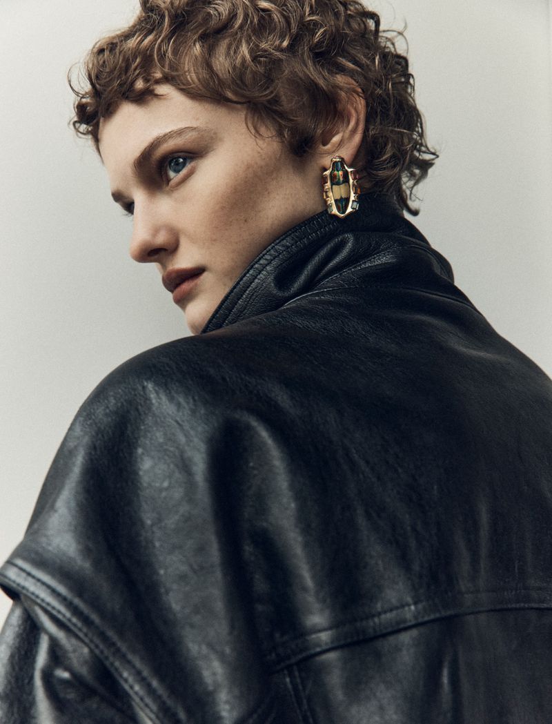 Woman with short curly hair looks over shoulder in studio wearing large gold statement earrings and a black leather jacket.