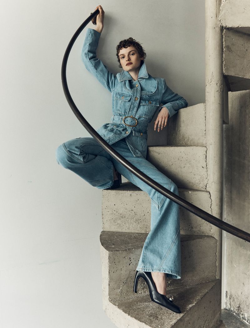 A young woman in a denim jacket and jeans poses on concrete stairs, holding onto a black metal railing, with black heels.