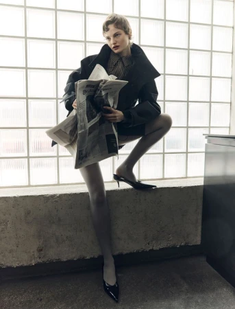 Stylish woman poses on concrete ledge in dark jacket, patterned top, gray tights, and heeled mules while holding newspapers.