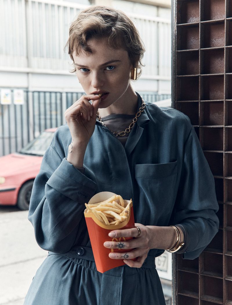 Young adult female in a blue jumpsuit eating fries from a red cardboard container in an urban setting next to a metal structure.