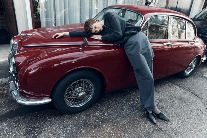 A person lounges on the hood of a vintage red car, posing in a relaxed manner, wearing grey casual clothing, outdoors in daylight.