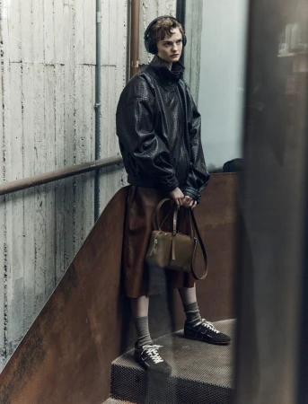Adult female model wearing an oversized black jacket and brown skirt stands on metal stairs with a leather purse and headphones.
