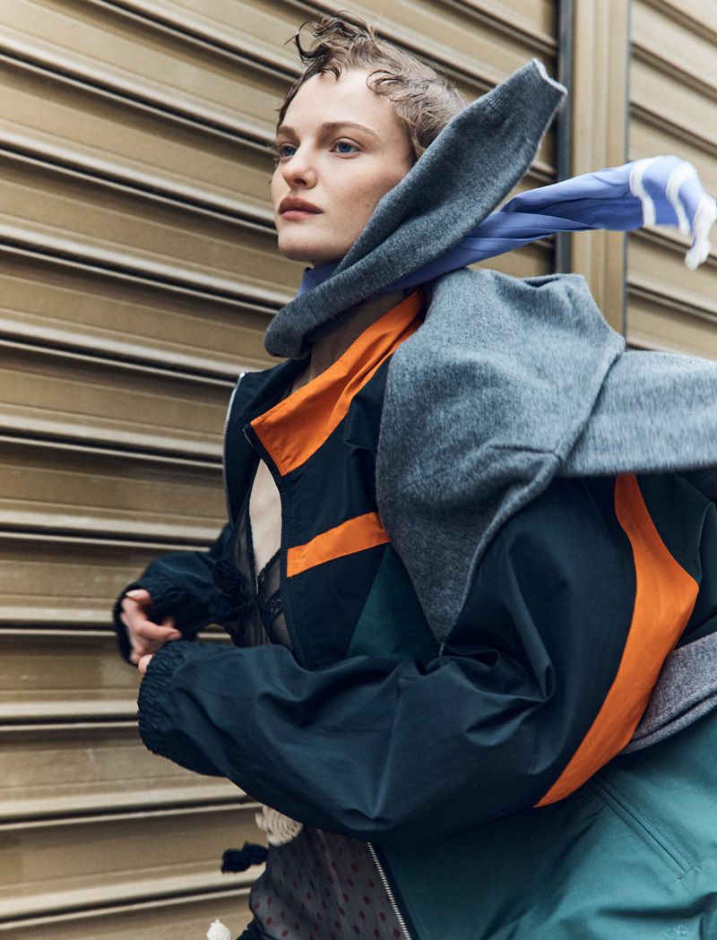 Woman in a dark jacket and grey scarf stands against a metal door, looking off to the side in an outdoor setting.