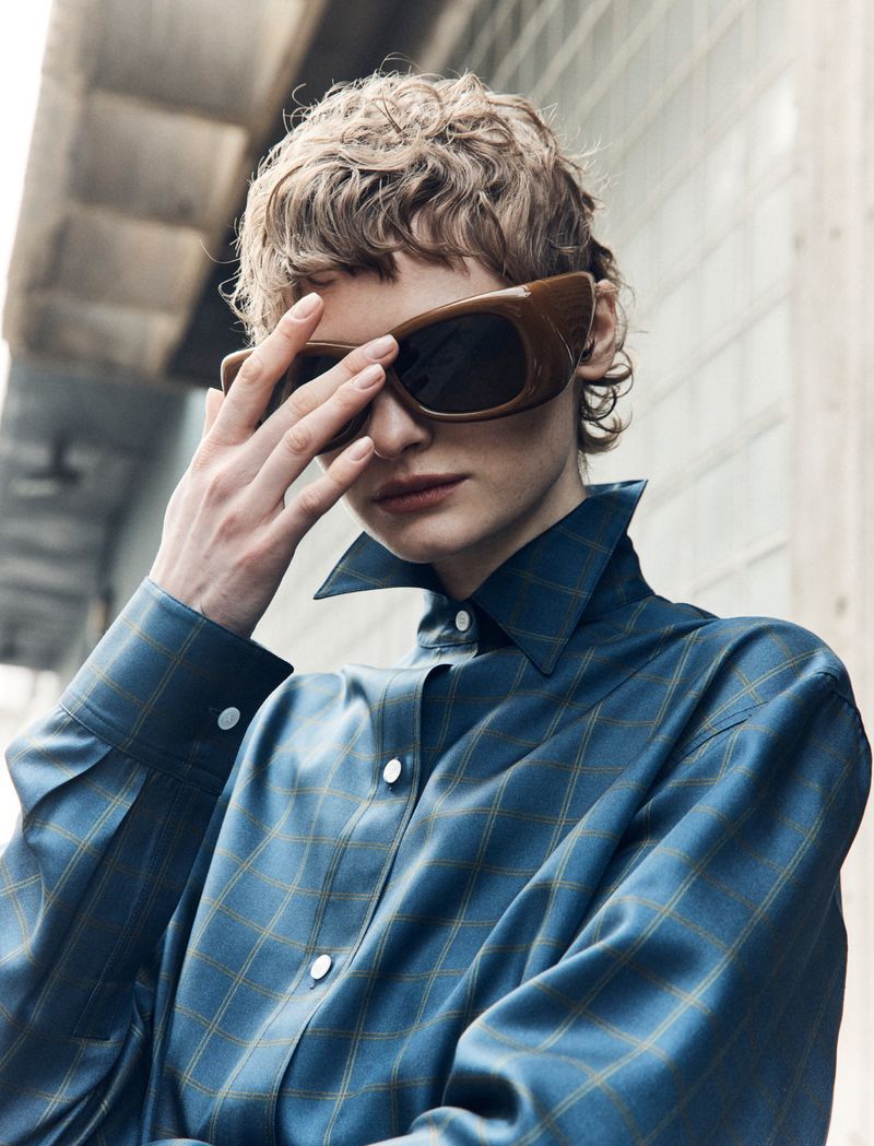 Brunette woman with short wavy hair poses in a teal and gold plaid shirt and oversized sunglasses against a neutral background.