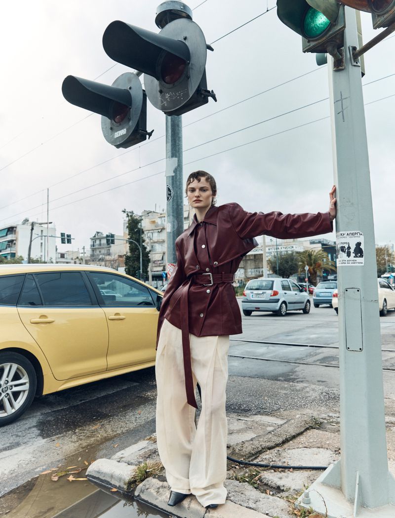 A fashionable adult woman posing in a maroon jacket and white pants, stands in front of a traffic light in an urban setting.
