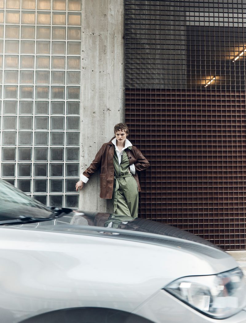 A person in an olive jumpsuit and brown jacket leans against a concrete wall next to a glass brick wall, with a silver car in the foreground.