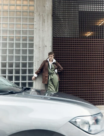 A person in an olive jumpsuit and brown jacket leans against a concrete wall next to a glass brick wall, with a silver car in the foreground.