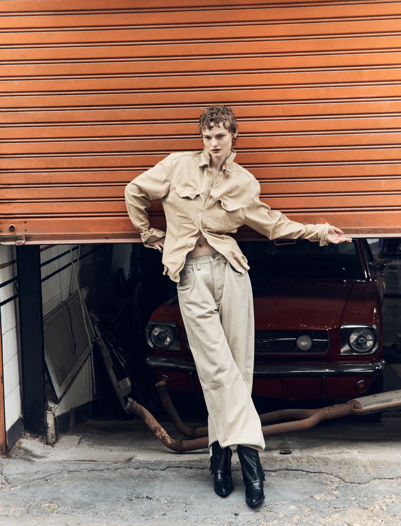 Adult female model poses leaning against orange roller door wearing beige jacket and pants with a burgundy vintage car in background.