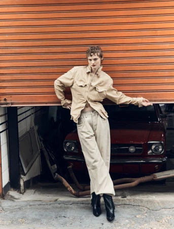 Adult female model poses leaning against orange roller door wearing beige jacket and pants with a burgundy vintage car in background.