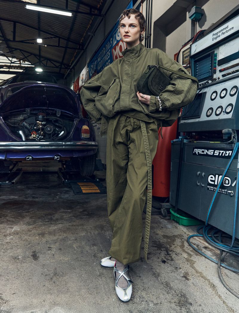 Fashion editorial shot of a woman in an olive-green tracksuit holding a purse, posed in an automotive repair shop in front of a purple car.
