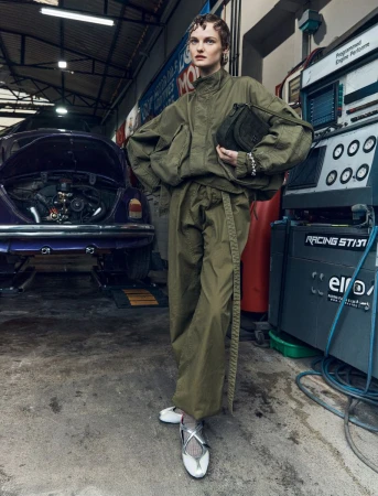 Fashion editorial shot of a woman in an olive-green tracksuit holding a purse, posed in an automotive repair shop in front of a purple car.