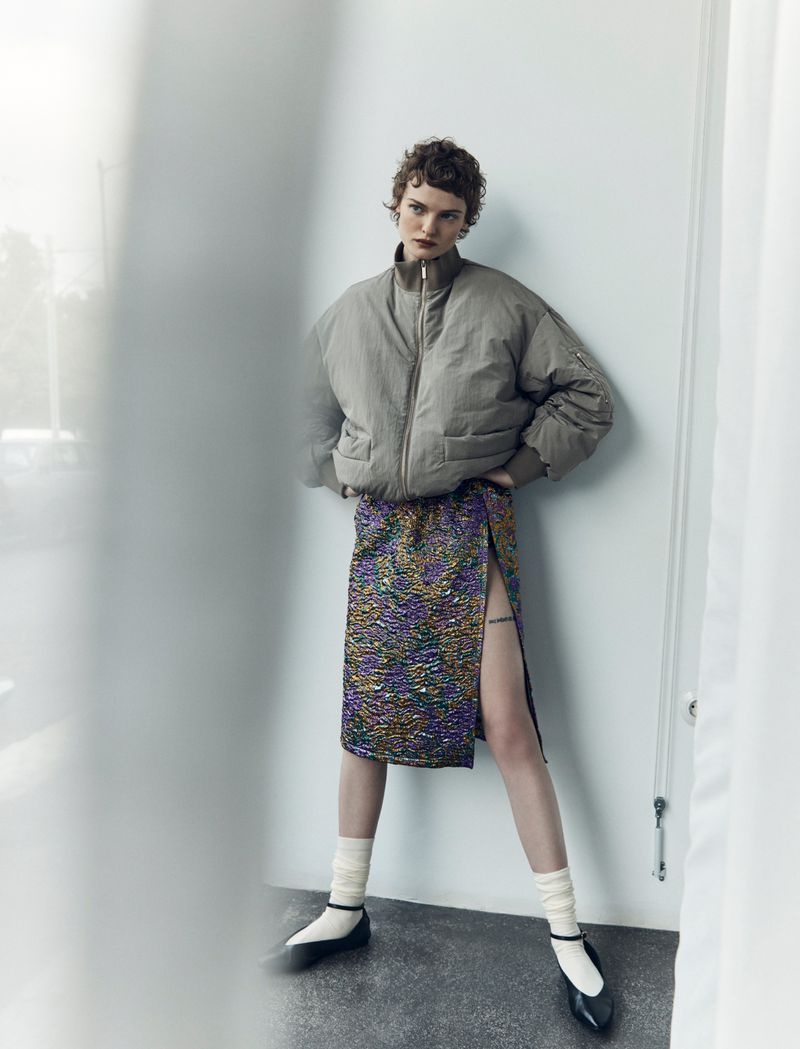 A stylish young woman poses indoors, wearing a taupe bomber jacket and a shimmering slit skirt, complemented by white socks and black flats.