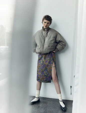 A stylish young woman poses indoors, wearing a taupe bomber jacket and a shimmering slit skirt, complemented by white socks and black flats.