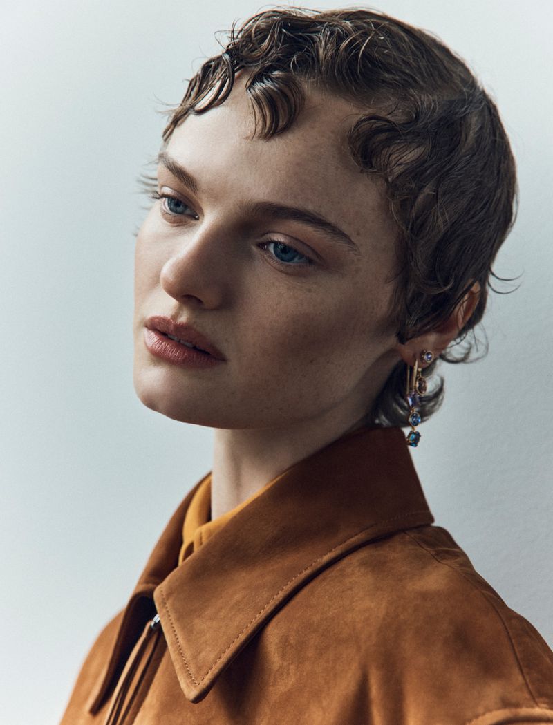 Portrait of a young adult female with short, dark wavy hair, wearing a brown suede jacket, looking off to the side in a neutral studio.