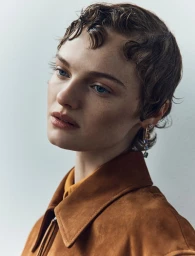 Close-up portrait of a woman with short wet-styled hair wearing a brown suede jacket against a plain light background