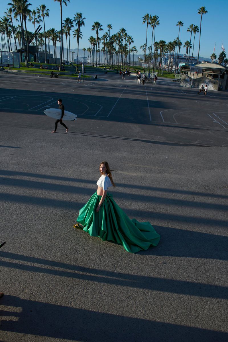 A woman in a green gown stands on an asphalt surface with long shadows, with palm trees and other people in the background.