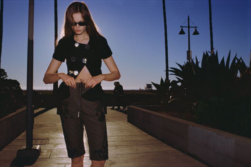 A slender adult woman wearing fashionable shorts and a top with embellishments, standing on a paved walkway at a beach.